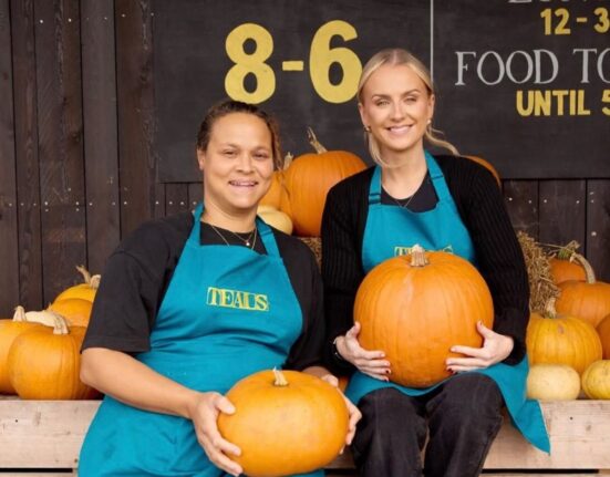 Two smiling members of staff at Teals Farm Shop, wearing teal aprons with the Teals logo, sit on wooden crates holding pumpkins in front of the farm shop’s opening hours sign