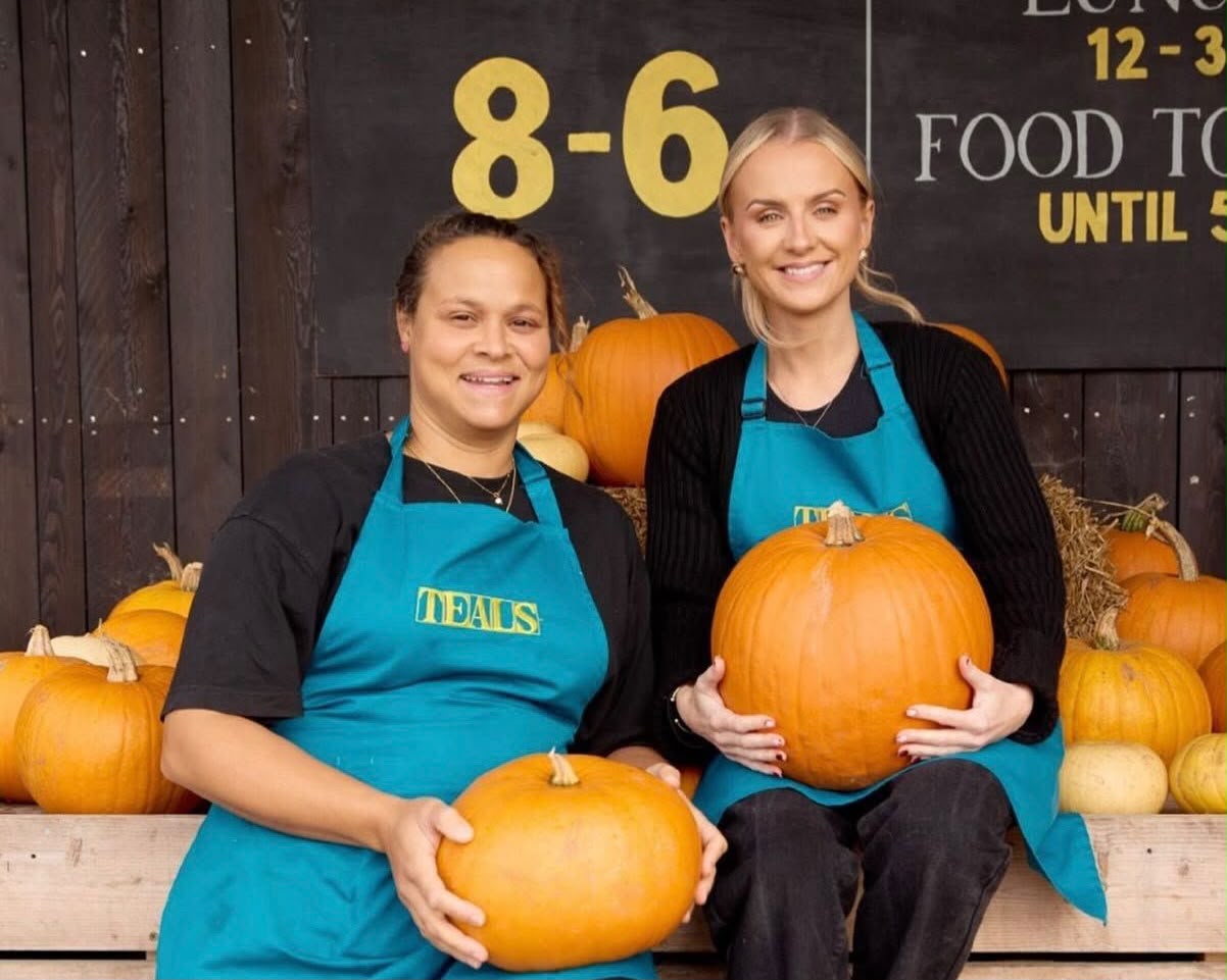 Two smiling members of staff at Teals Farm Shop, wearing teal aprons with the Teals logo, sit on wooden crates holding pumpkins in front of the farm shop’s opening hours sign