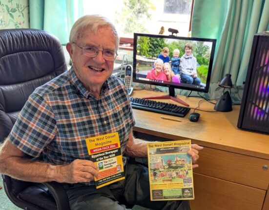 Bob Holman, 82, of West Dorset Magazine, working past retirement age, smiles at his desk holding a copy of the magazine and its 2026 media pack.