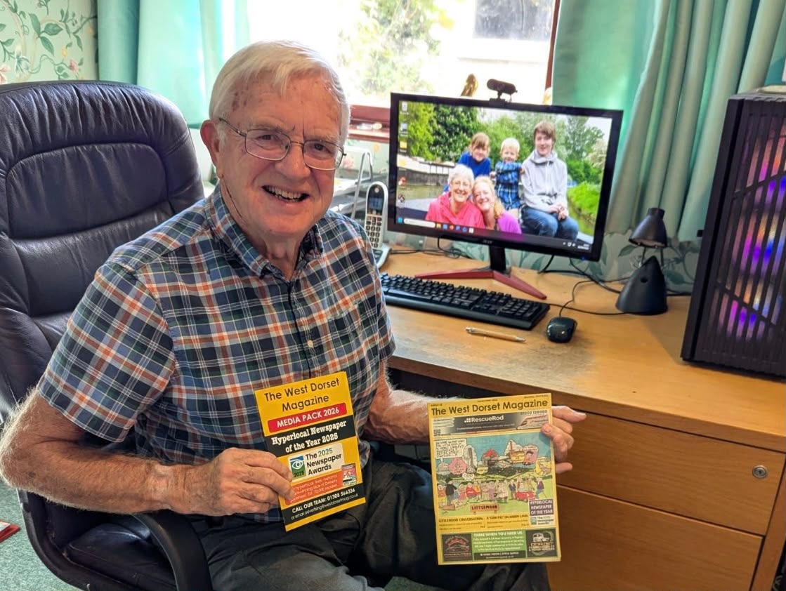 Bob Holman, 82, of West Dorset Magazine, working past retirement age, smiles at his desk holding a copy of the magazine and its 2026 media pack.