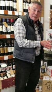Martin Ridley, 80, who runs St Michael’s Trading Estate in Bridport, working past retirement age, holds a glass among shelves of wine bottles.