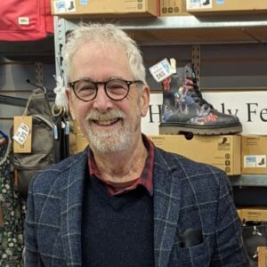 Riff Tilbury, 76, of Steptoes shoe shop in Bridport, working past retirement age, stands smiling among shelves of boots and shoe boxes.