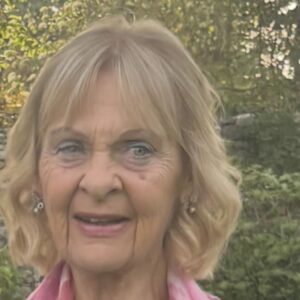 Anne Roots, 76, of Nutley Farm, working past retirement age, smiles outdoors in a pink top against a leafy background.